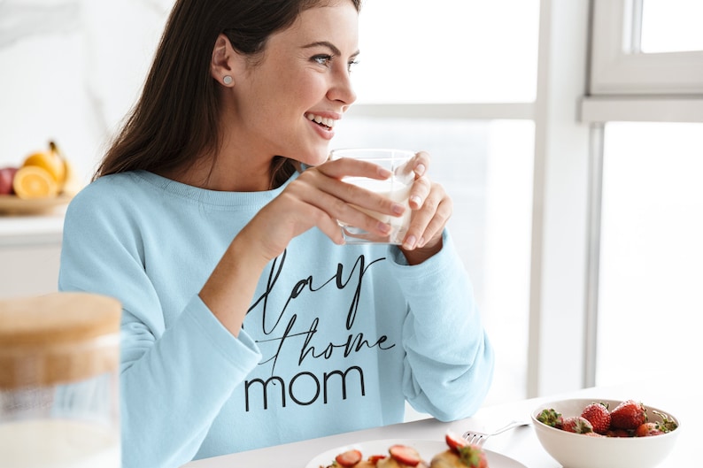 May include: A woman wearing a light blue sweatshirt with the text "Stay Home Mom" in black lettering. She is holding a glass of milk and smiling.