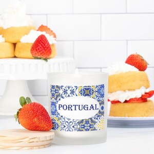 May include: A white frosted glass candle with a blue and yellow patterned label that says "PORTUGAL". The candle is sitting on a white surface with a strawberry and a wooden coaster.