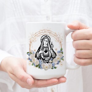 May include: White ceramic mug with a black and white image of the Virgin Mary with a heart in her chest. The image is surrounded by a gold floral wreath with white and blue flowers.