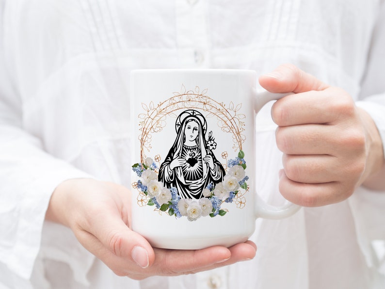 May include: White ceramic mug with a black and white image of the Virgin Mary with a heart in her chest. The image is surrounded by a gold floral wreath with white and blue flowers.
