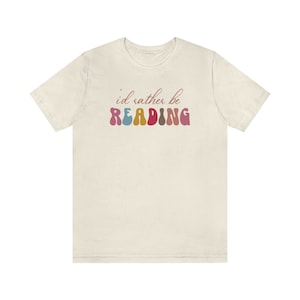 Puede incluir: Una camiseta beige claro con el texto "i'd rather be READING" en letras de colores onduladas.