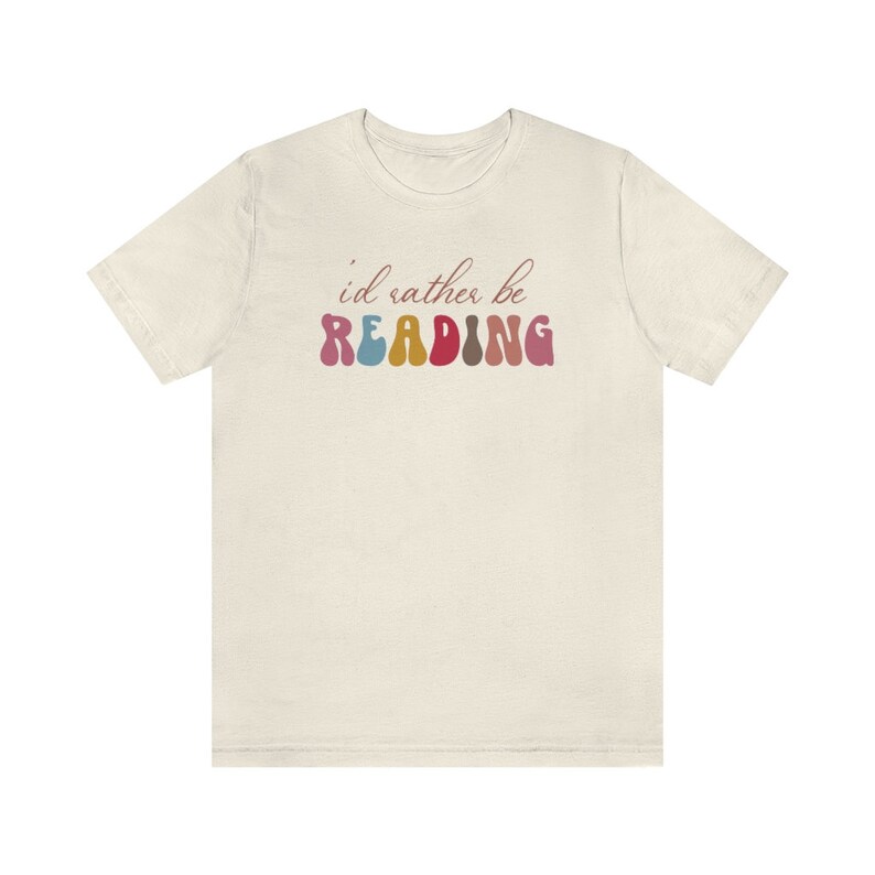 Puede incluir: Una camiseta beige claro con el texto "i'd rather be READING" en letras de colores onduladas.