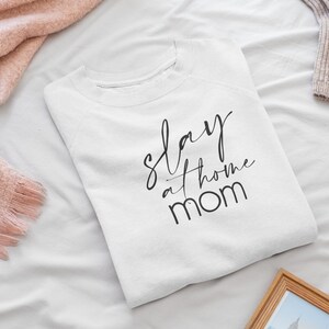 May include: White t-shirt with black text that reads "slay at home mom".