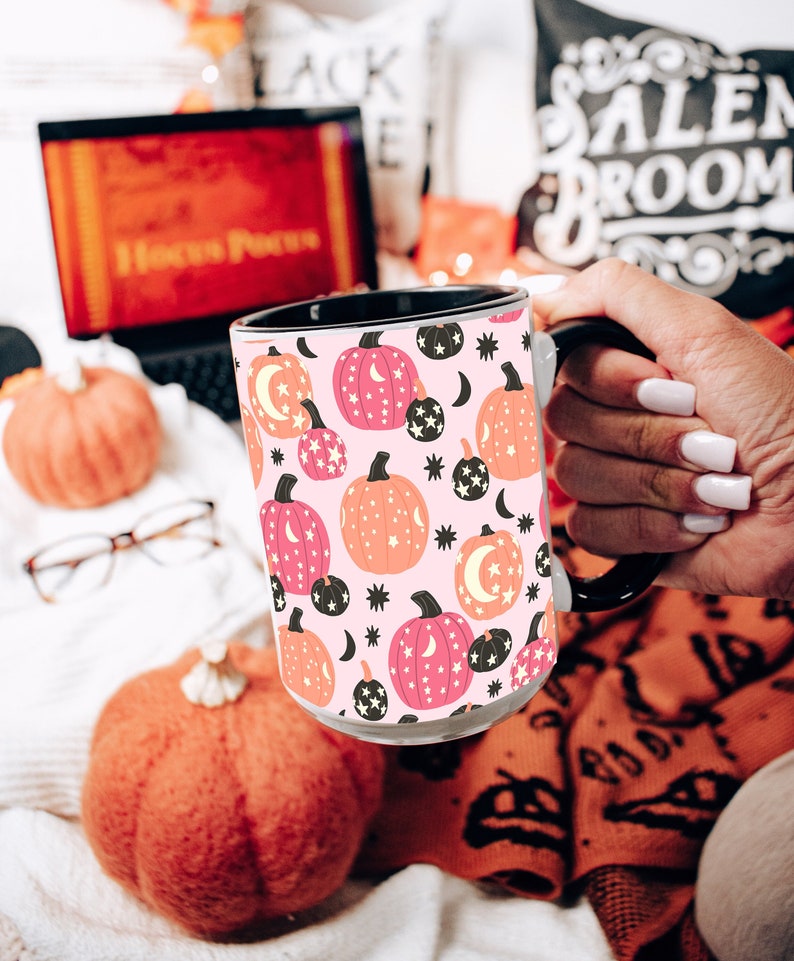 May include: A white ceramic mug with a black handle features a pink background with a repeating pattern of orange pumpkins with pink stars and black crescent moons.