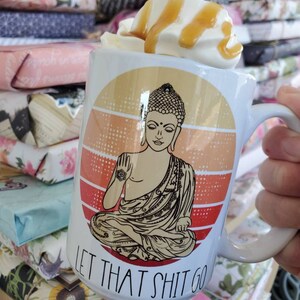 K&ouml;nnte beinhalten: Wei&szlig;e Keramiktasse mit einem Retro-Sonnenuntergangsdesign, das einen meditierenden Buddha und den Text "Let That Shit Go" zeigt. Die Tasse ist mit einem Kaffeegetr&auml;nk gef&uuml;llt, das mit Schlagsahne und Karamellsauce garniert ist.