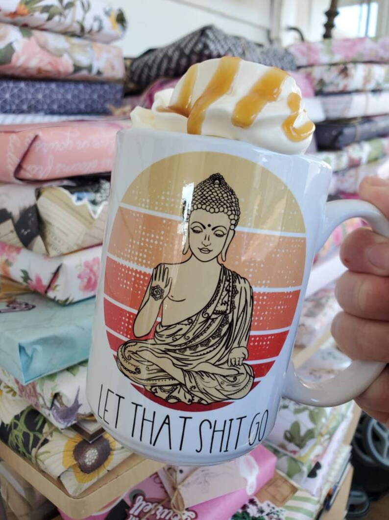 K&ouml;nnte beinhalten: Wei&szlig;e Keramiktasse mit einem Retro-Sonnenuntergangsdesign, das einen meditierenden Buddha und den Text "Let That Shit Go" zeigt. Die Tasse ist mit einem Kaffeegetr&auml;nk gef&uuml;llt, das mit Schlagsahne und Karamellsauce garniert ist.