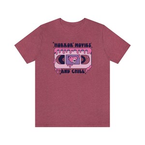 May include: A heather red t-shirt with a graphic of a pink VHS tape with the text "HORROR MOVIES AND CHILL" in a retro font.