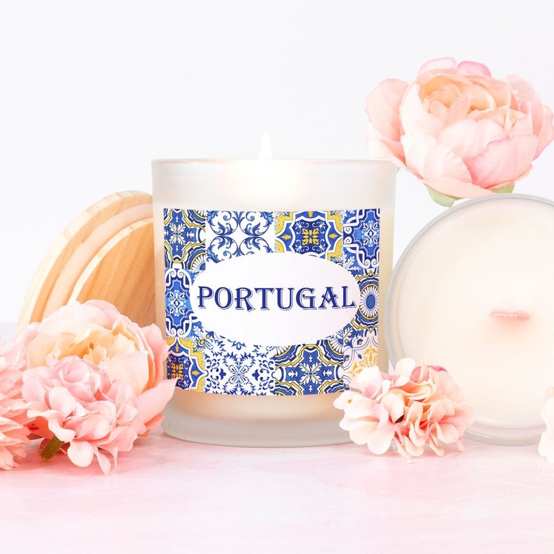 May include: A white candle with a blue and yellow patterned label that says "PORTUGAL". The candle is in a glass jar with a wooden lid. The candle is surrounded by pink flowers.