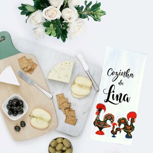 May include: A white kitchen towel with the text "Cozinha da Lina" in black script and two colorful rooster illustrations. The towel is on a white surface with a cheese board, crackers, apples, and olives.