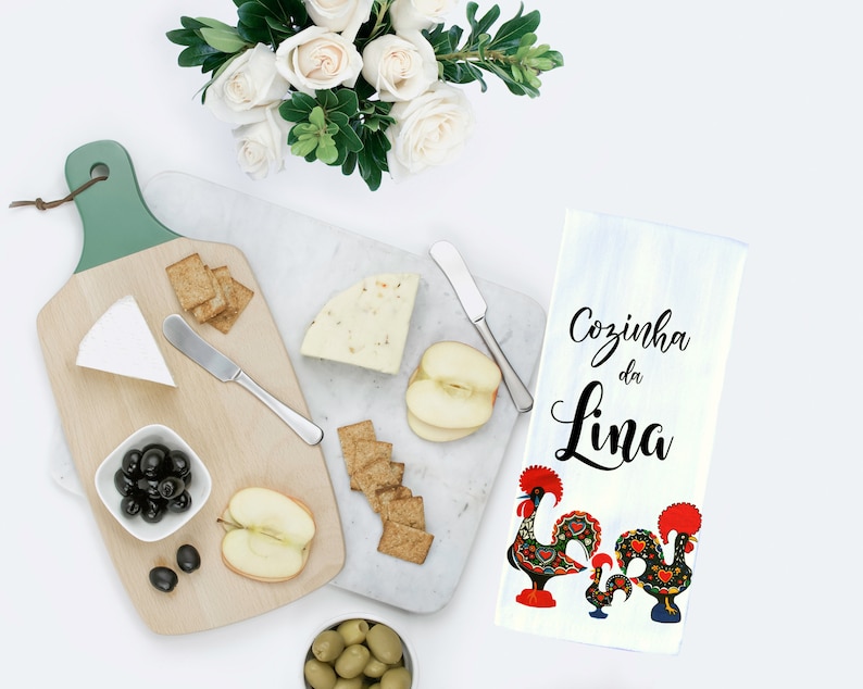 May include: A white kitchen towel with the text "Cozinha da Lina" in black script and two colorful rooster illustrations. The towel is on a white surface with a cheese board, crackers, apples, and olives.