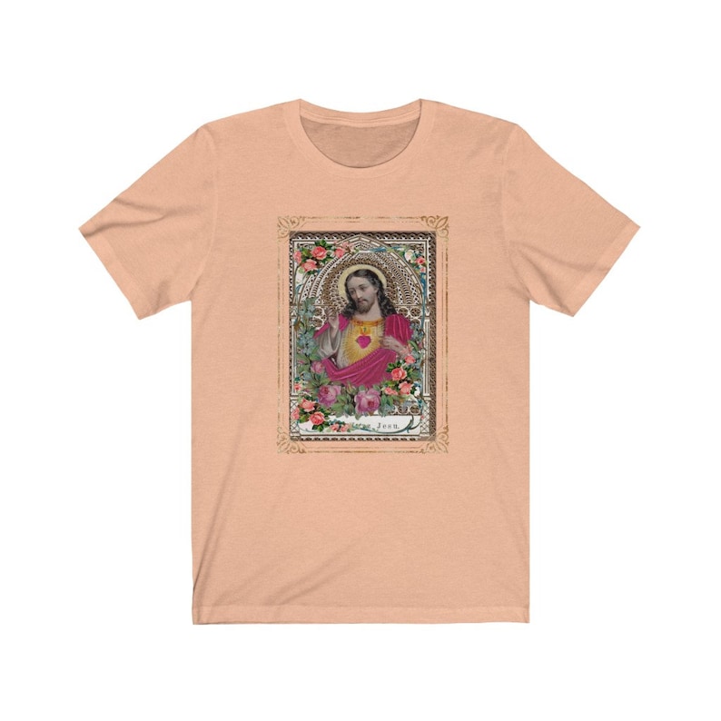 May include: A peach-colored t-shirt with a vintage-style image of Jesus in a gold frame with floral accents. The image is surrounded by a gold frame with ornate details. The text "Jesus" is printed below the image.