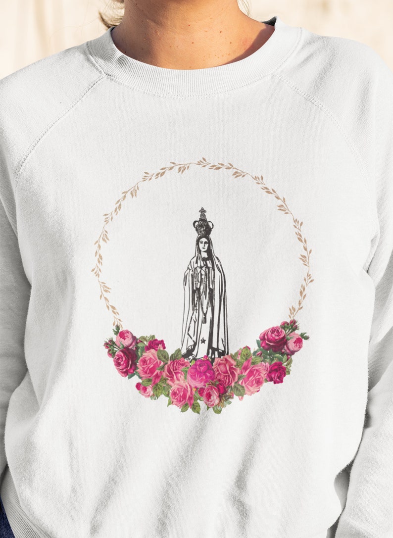 Pu&ograve; includere: Felpa bianca con un'illustrazione in bianco e nero della Vergine Maria in una ghirlanda di foglie marroni e rose rosa.