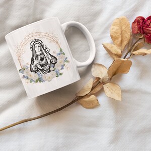 May include: White ceramic mug with a black and white image of the Virgin Mary with a heart and flowers. The mug is on a white surface with a dried rose.