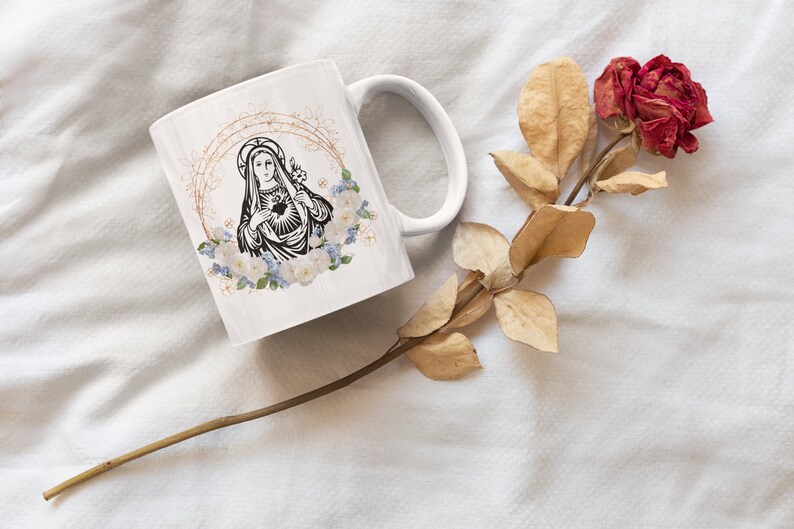 May include: White ceramic mug with a black and white image of the Virgin Mary with a heart and flowers. The mug is on a white surface with a dried rose.