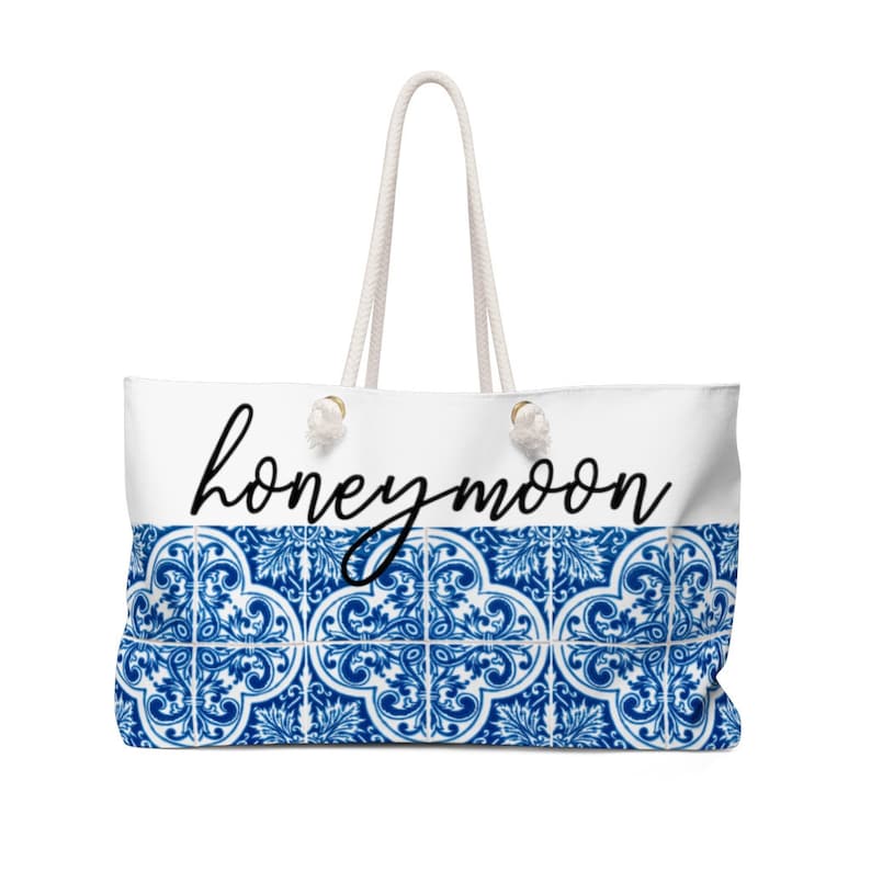 Op de afbeelding: Witte draagtas met touwhandvatten en een blauw-wit tegelpatroon aan de onderkant. Het woord "honeymoon" staat in zwart script over de bovenkant van de tas.