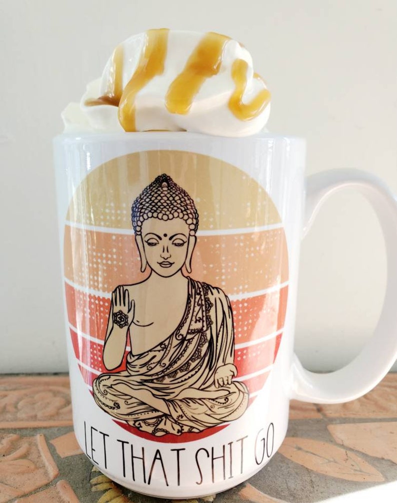 K&ouml;nnte beinhalten: Eine wei&szlig;e Keramiktasse mit einer Grafik eines meditierenden Buddha in einem kreisf&ouml;rmigen Sonnenuntergangsmuster. Der Text "Let That Shit Go" ist unter dem Buddha aufgedruckt. Die Tasse hat einen wei&szlig;en Henkel und ist mit Schlagsahne und Karamellso&szlig;e verziert.
