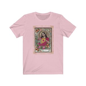 May include: A pink t-shirt with a vintage-style image of Jesus in a gold frame. The image features Jesus with a heart in his hand and roses surrounding him. The text "Jesus" is at the bottom of the image.