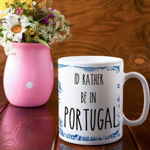 May include: White ceramic mug with a blue and white patterned border. The mug has the text "I'd rather be in Portugal" printed in black.