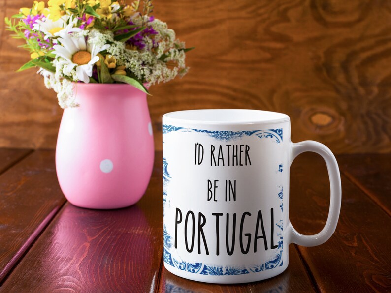 May include: White ceramic mug with a blue and white patterned border. The mug has the text "I'd rather be in Portugal" printed in black.