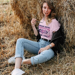 May include: A woman wearing a pink t-shirt with a cassette tape graphic and the text "Horror Movies and Chill". She is sitting in a field of hay with a black leather jacket and blue jeans.