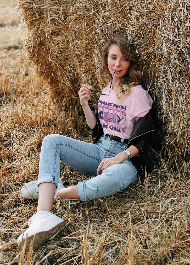 May include: A woman wearing a pink t-shirt with a cassette tape graphic and the text "Horror Movies and Chill". She is sitting in a field of hay with a black leather jacket and blue jeans.
