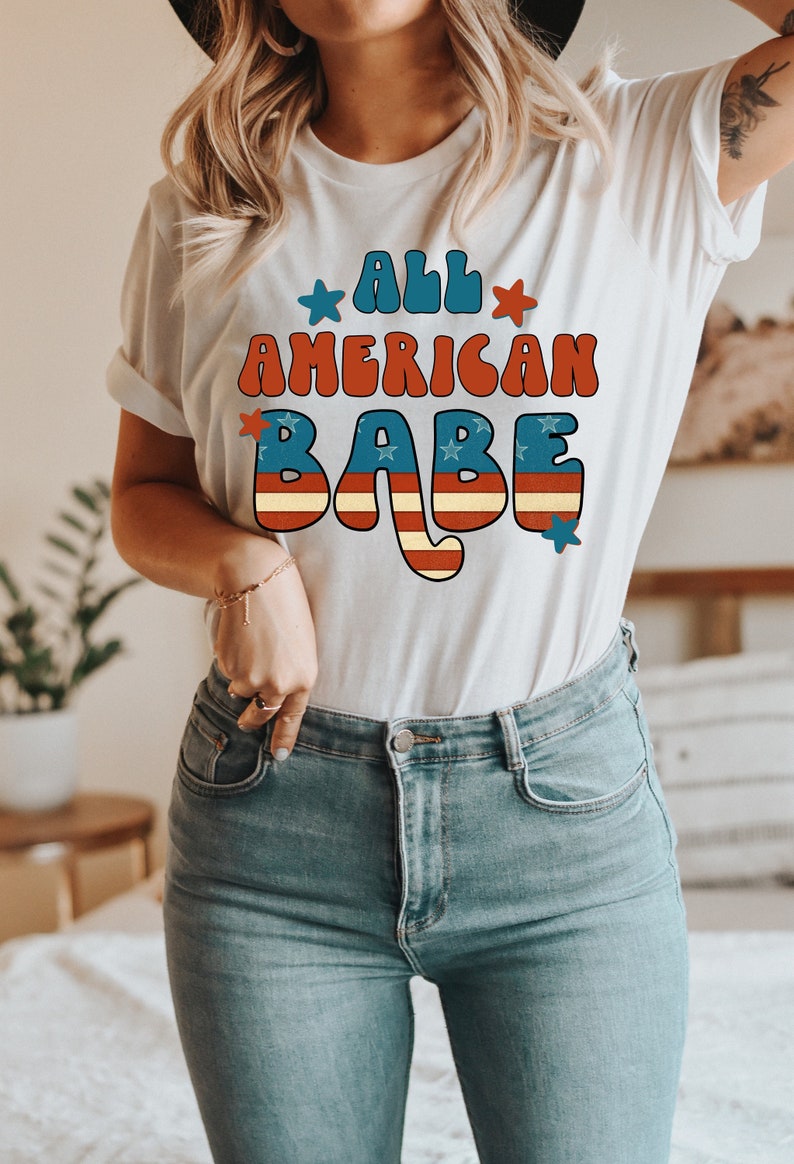 May include: A white t-shirt with a red, white and blue graphic that says "All American Babe" in a retro font.