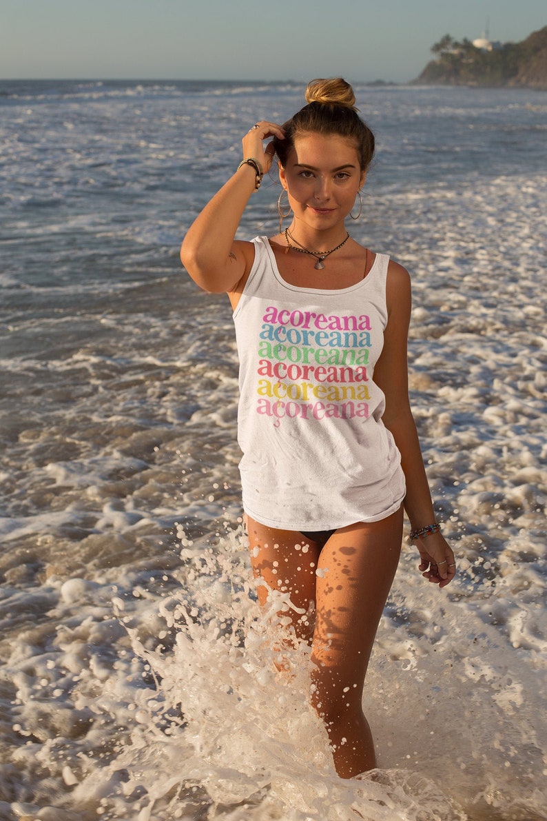 Op de afbeelding: Een vrouw draagt een wit tanktop met het woord "acoreana" dat in regenboogkleuren wordt herhaald. Ze staat in de oceaan met golven die om haar heen breken.
