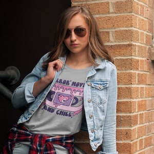 May include: A woman wearing a light gray t-shirt with a pink and purple graphic of a cassette tape with the text "Horror Movie and Chill". She is also wearing a blue denim jacket and a red and black plaid shirt tied around her waist.