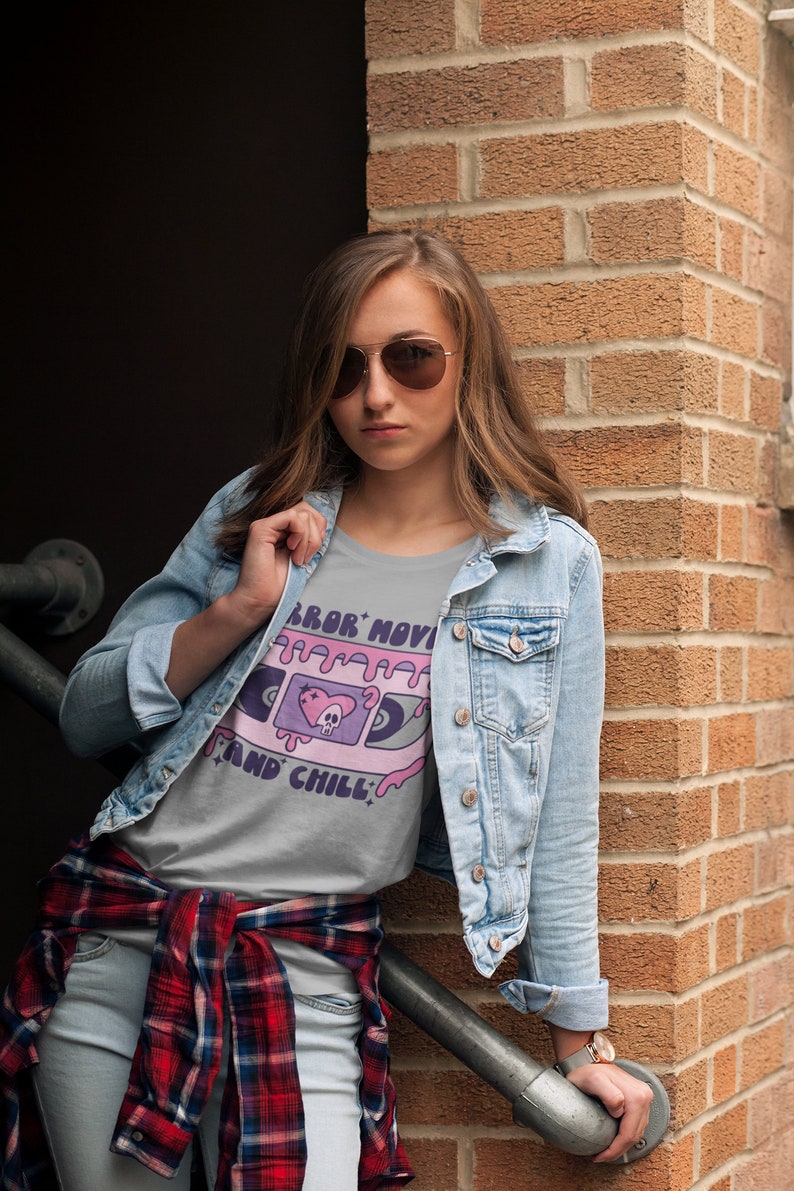 May include: A woman wearing a light gray t-shirt with a pink and purple graphic of a cassette tape with the text "Horror Movie and Chill". She is also wearing a blue denim jacket and a red and black plaid shirt tied around her waist.
