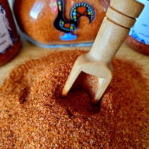 May include: A close-up of a wooden scoop filled with a bright orange-red spice powder. The powder is piled high and the scoop is resting on a wooden surface. A glass jar with a red rooster design is visible in the background.
