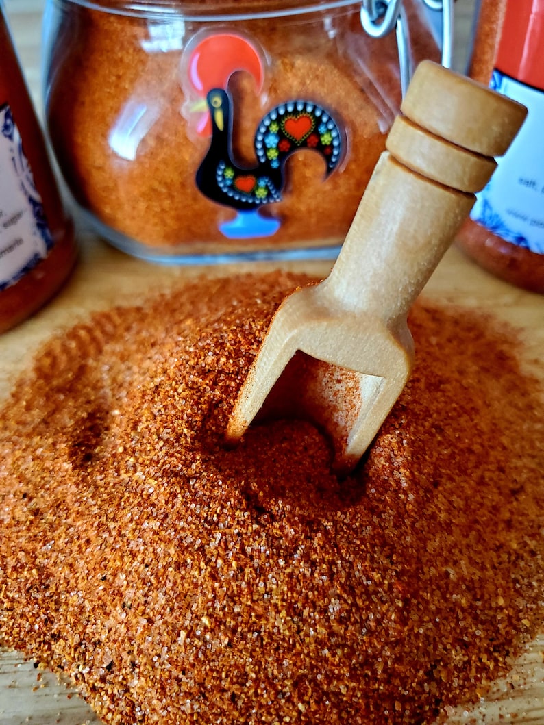 May include: A close-up of a wooden scoop filled with a bright orange-red spice powder. The powder is piled high and the scoop is resting on a wooden surface. A glass jar with a red rooster design is visible in the background.