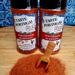 May include: Two glass jars of red Portuguese seasoning with black lids and white labels. The labels say "Taste Portugal" and "Tia Maria's Portuguese Seasoning". The jars are on a wooden surface with a small wooden scoop.