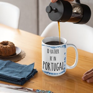 May include: White ceramic mug with a blue and white patterned design. The mug has the text "I'd rather be in Portugal" printed on it. A person is pouring coffee from a French press into the mug.