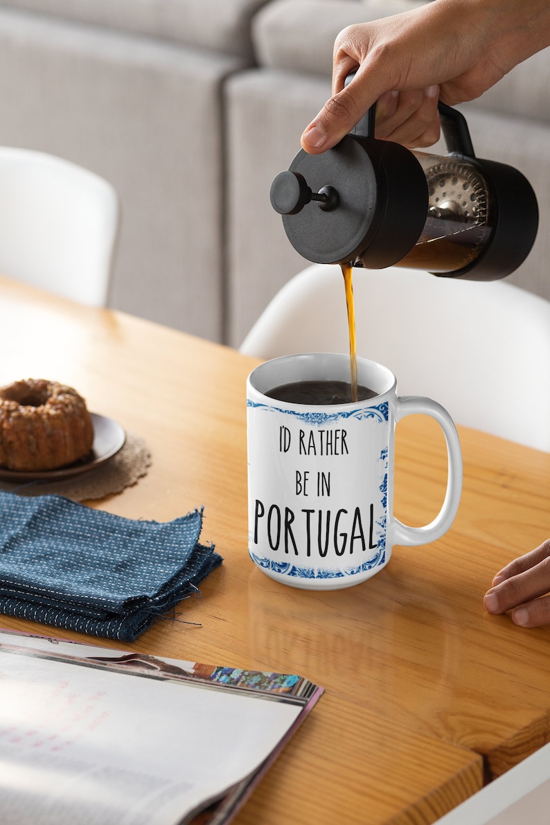May include: White ceramic mug with a blue and white patterned design. The mug has the text "I'd rather be in Portugal" printed on it. A person is pouring coffee from a French press into the mug.