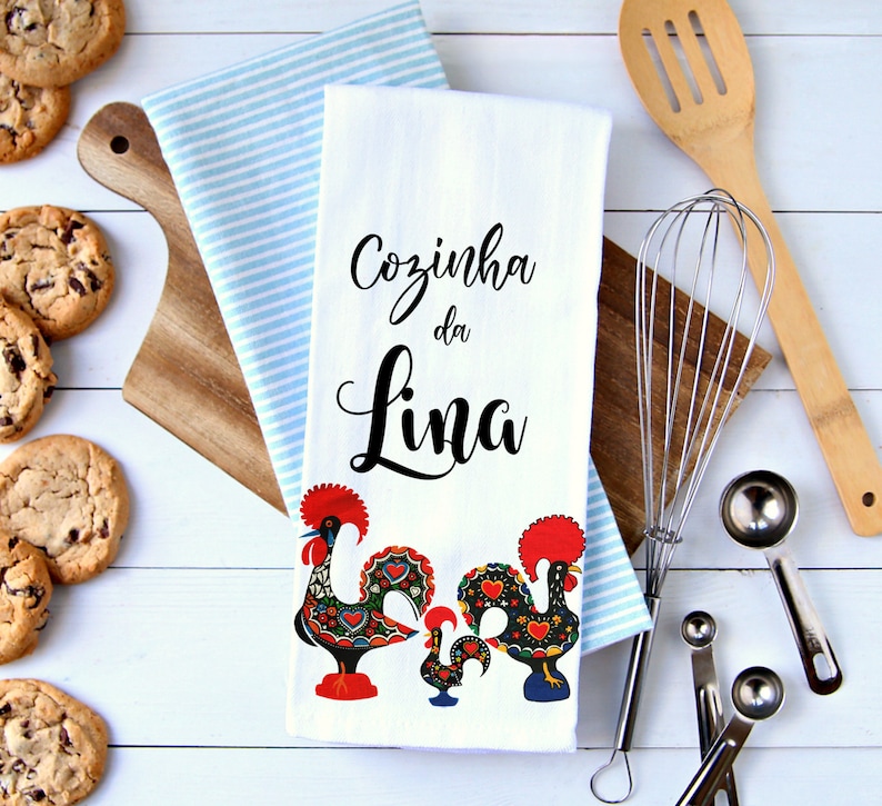 May include: White kitchen towel with the text "Cozinha da Lina" in black cursive lettering. Two colorful rooster illustrations are at the bottom of the towel.
