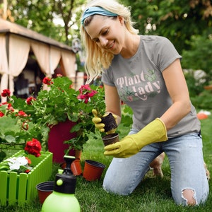 May include: A woman wearing a grey t-shirt with the text "Plant Lady" in pink lettering and a green cactus graphic is kneeling in a garden. She is holding a small potted plant and wearing yellow gardening gloves.