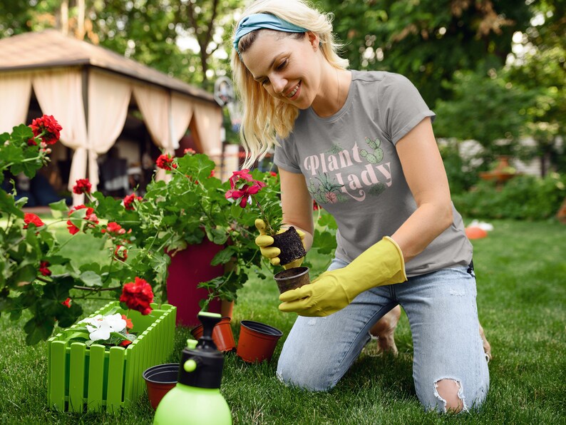 May include: A woman wearing a grey t-shirt with the text "Plant Lady" in pink lettering and a green cactus graphic is kneeling in a garden. She is holding a small potted plant and wearing yellow gardening gloves.