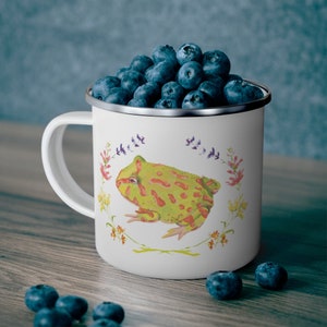 May include: White enamel mug with a green and red frog illustration surrounded by a floral wreath. The mug has a white handle and is filled with blueberries.