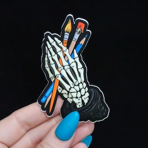 May include: A sticker featuring a skeletal hand holding three paintbrushes. The brushes are orange, blue, and red. The hand is wearing a black glove. The sticker is on a black background.