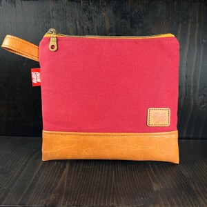 May include: A red canvas zippered pouch with a brown leather base and a small brown leather patch with a logo. The pouch has a brown leather strap.