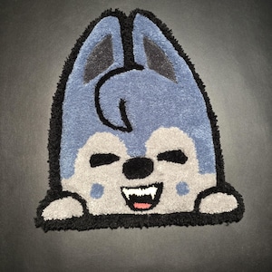 May include: A handmade rug in the shape of a cartoon wolf, featuring a smiling face with a black nose, white teeth, and a pink tongue. The wolf has blue and gray fur with black outlines and accents. The rug is on a dark surface.