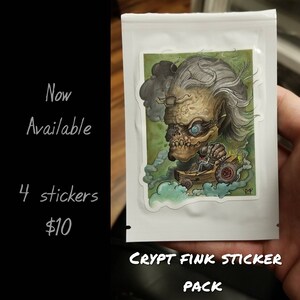May include: A sticker pack featuring a cartoon illustration of a green-skinned, gray-haired creature with one blue eye driving a small, wooden car. The sticker pack is labeled "CRYPT FINK STICKER PACK".