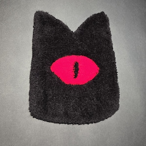 May include: A black, plush cat-shaped rug with a vibrant red eye design. The rug has a soft, textured appearance and a unique, eye-catching design. This decorative rug is perfect for a themed room.