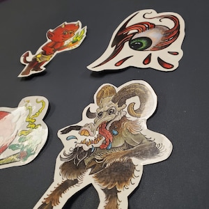 May include: Four vintage tattoo flash designs featuring a red devil holding a torch, a green eye with red and black accents, a horned creature with a long tongue, and a pink and green floral design.