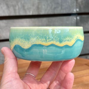 Puede incluir: Un pequeño cuenco de cerámica azul verdoso con un glaseado amarillo alrededor del borde. El cuenco está hecho a mano y tiene un diseño ondulado único.