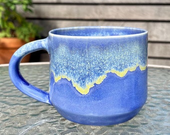 May include: A blue ceramic mug with a yellow glaze design. The mug has a rounded handle and a wide base.