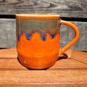 May include: A handmade ceramic mug with a vibrant orange base and handle. The upper portion transitions from a dark teal to a lighter orange, with blue accents. The mug is designed for hot beverages.