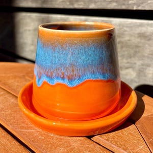 Può includere: Un vaso in ceramica con piattino abbinato, caratterizzato da una base smaltata color arancione vivace che sfuma in uno smalto blu testurizzato e colante vicino al bordo. Il contenitore cilindrico ha una finitura liscia e un'ampia apertura, che mette in risalto un netto contrasto tra i toni arancione brillante e blu intenso.