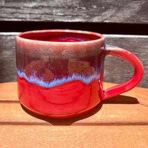 May include: A ceramic mug with a glossy finish, featuring a vibrant red base that transitions into a deep maroon upper section. A distinct, thin band of light blue glaze creates a textured, feathered effect where the red and maroon colors meet.