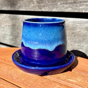 May include: A ceramic planter with a glossy finish featuring a deep royal blue base that transitions into a lighter sky blue glaze at the top rim. The planter sits securely on a matching circular blue saucer with a smooth, reflective surface.